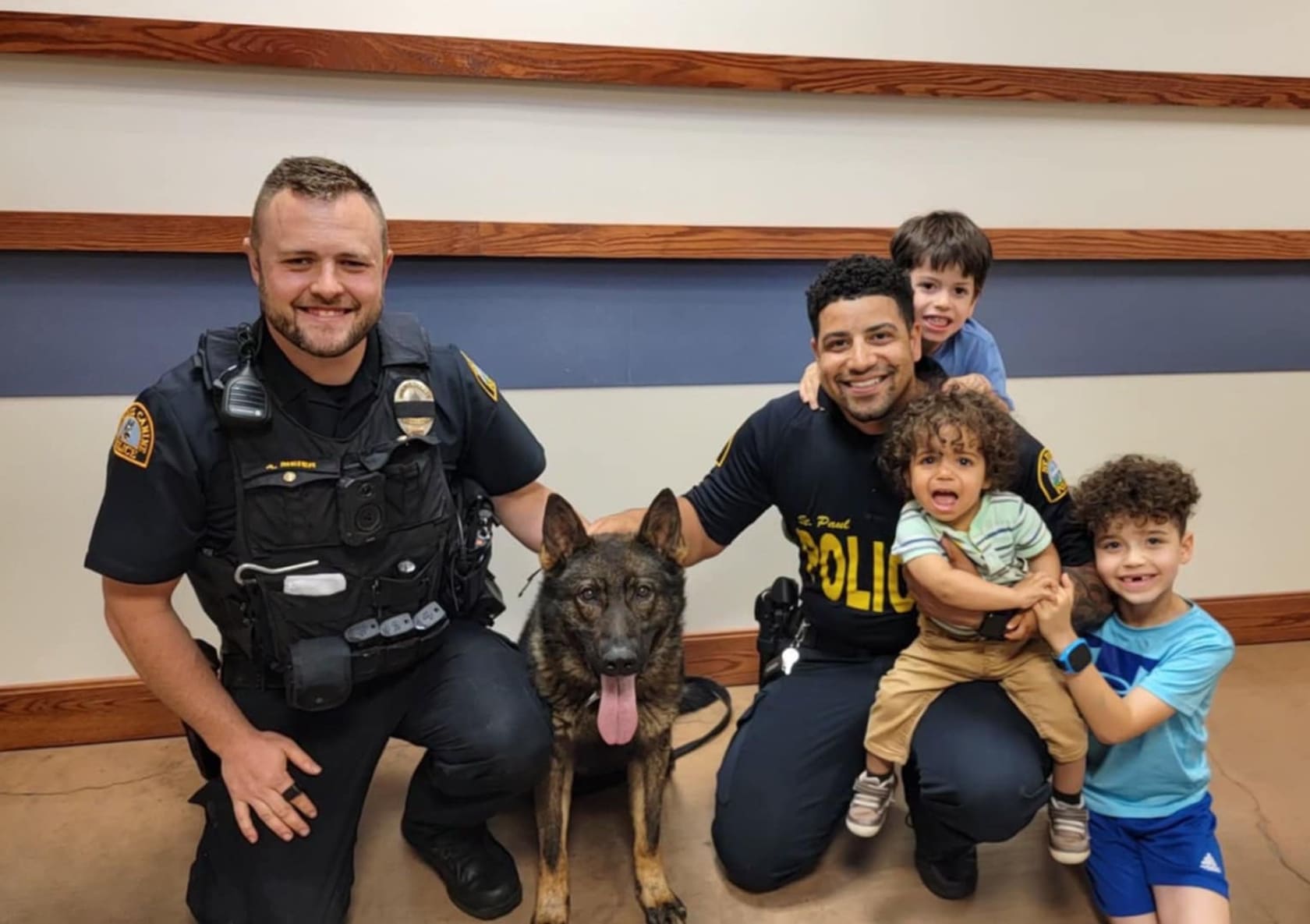 Two police officers kneeling beside a German Shepherd police dog with three children smiling around them indoors.