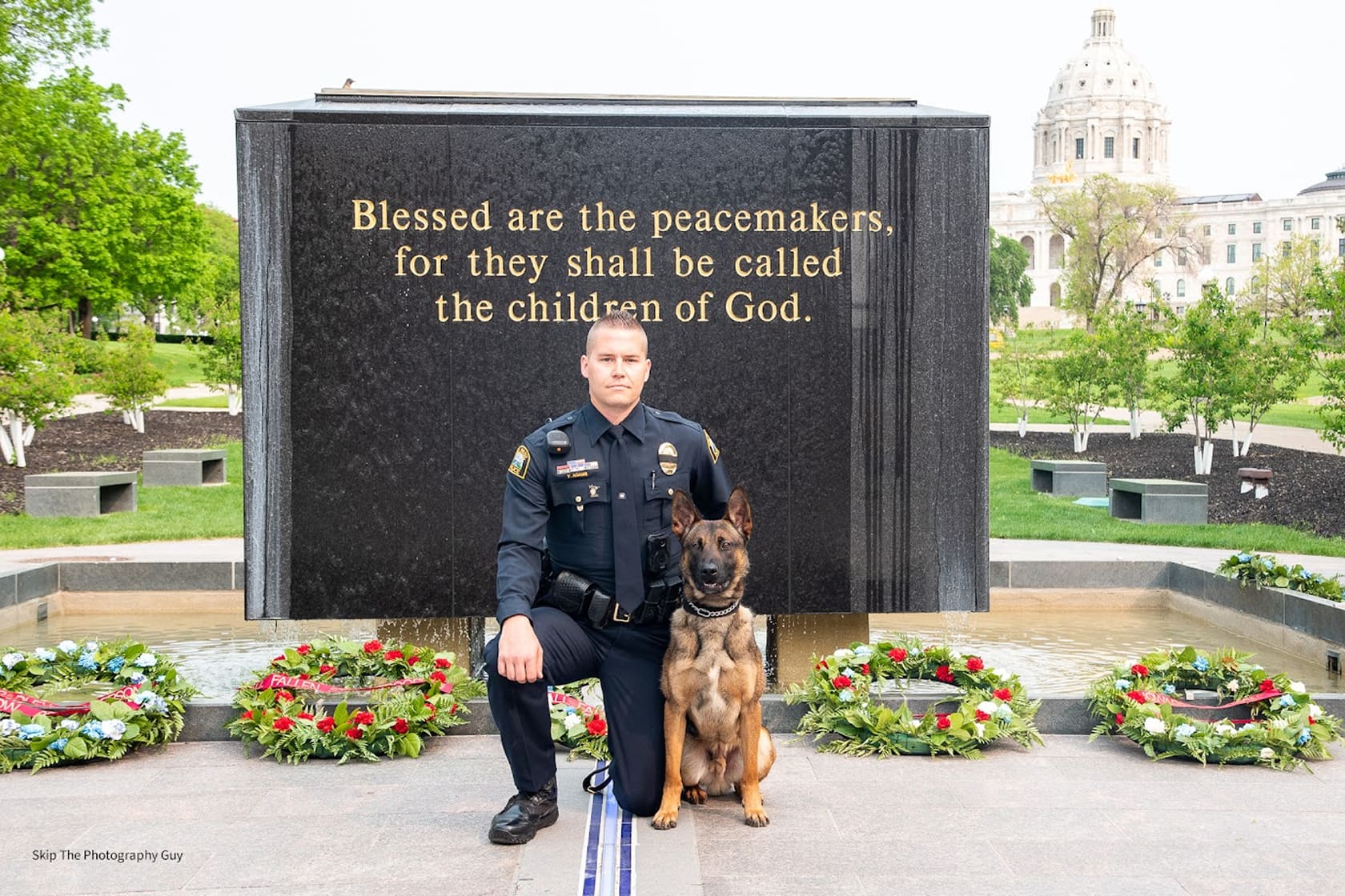 Police officer kneeling beside a German Shepherd in front of a memorial engraved with the quote 'Blessed are the peacemakers, for they shall be called the children of God.'