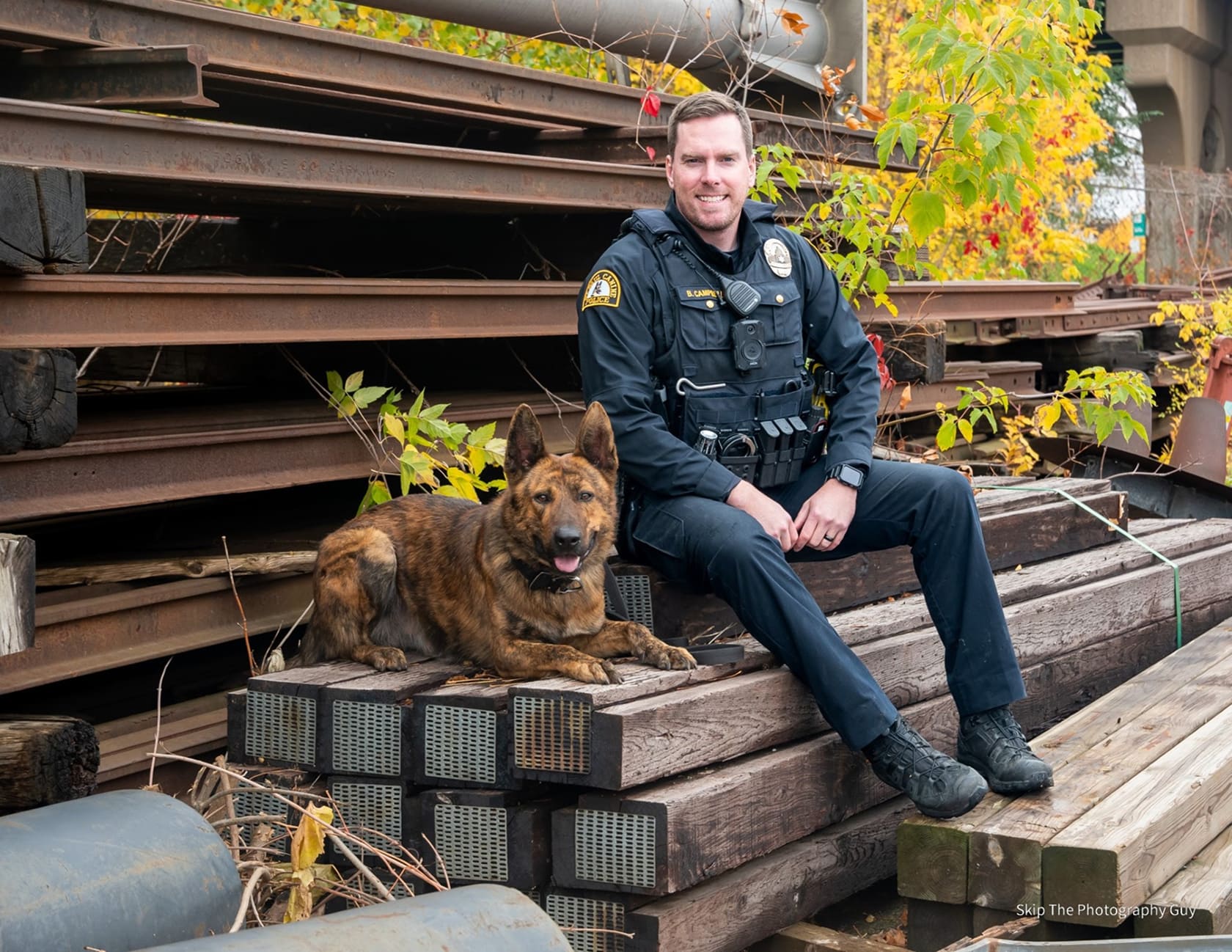 Police officer in uniform sitting on stacked wooden beams next to a brown police dog.