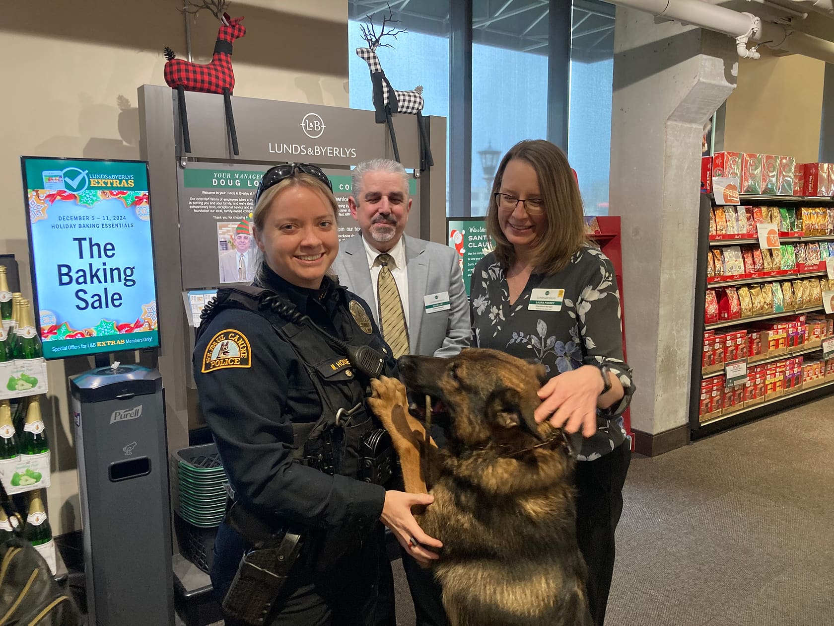 A female police officer and two store employees smile as they pet a large German Shepherd inside a grocery store near a sign for a holiday baking sale.