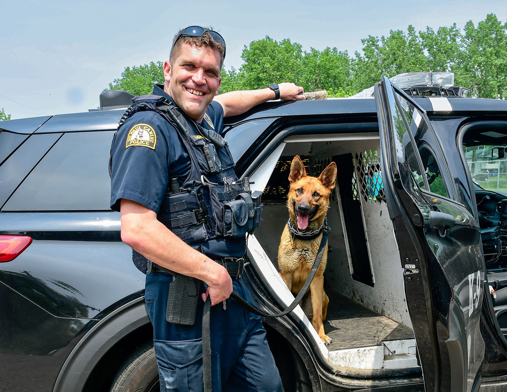 Smiling police officer standing beside a black K-9 unit vehicle with a German Shepherd dog inside.