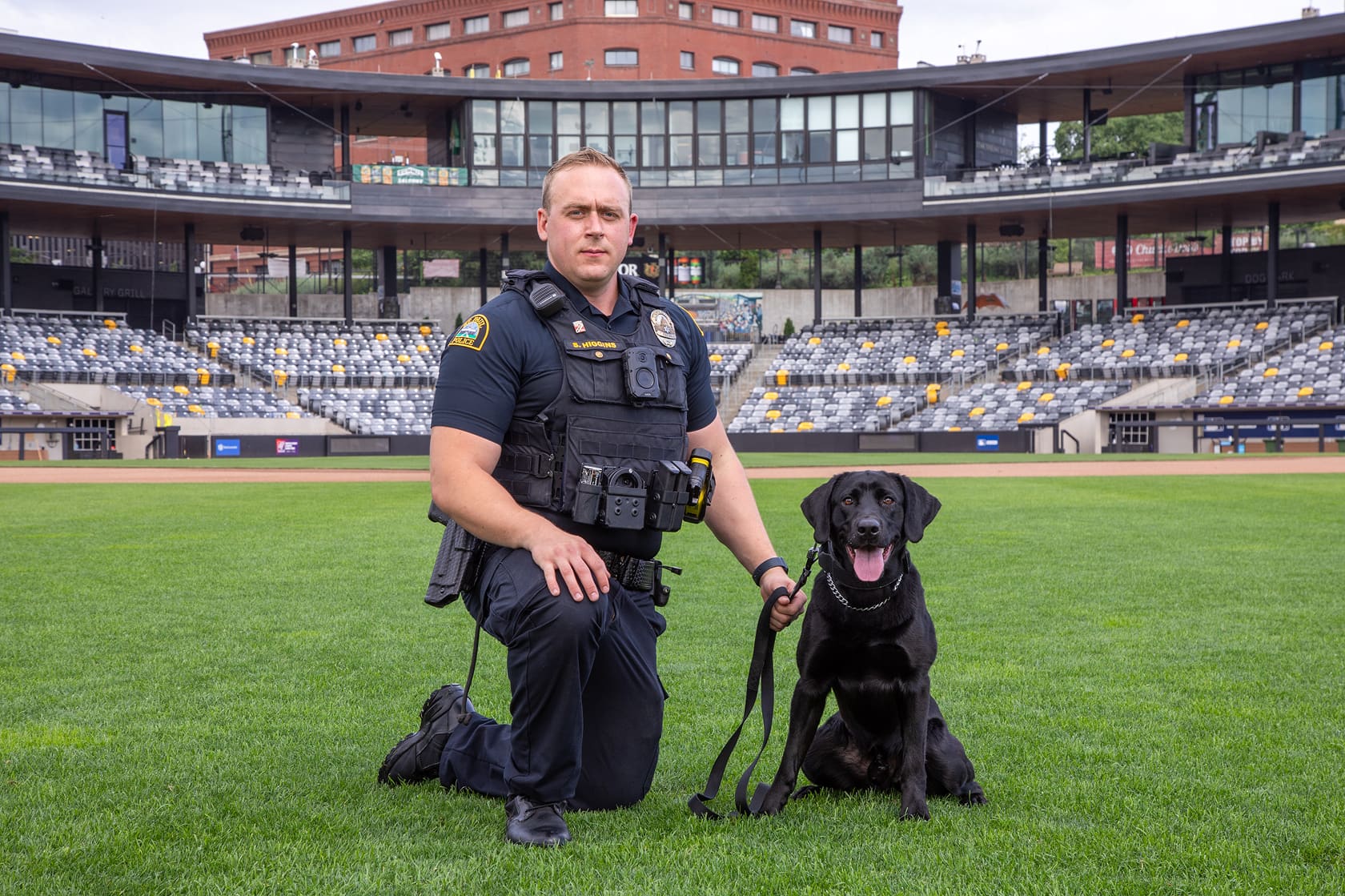 Police officer in uniform kneeling on grass with a black Labrador dog on a leash in an empty stadium.
