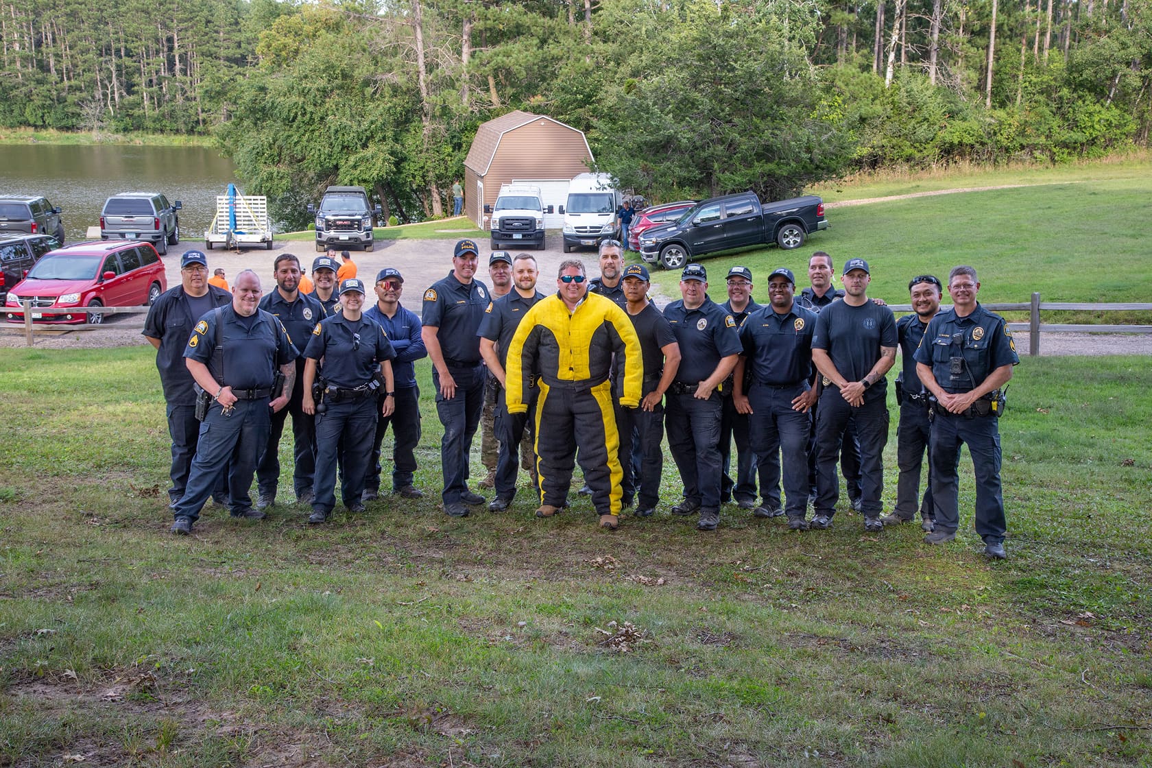 Group of police officers standing on grass with one person in a yellow and black bomb suit, with vehicles and trees in the background.
