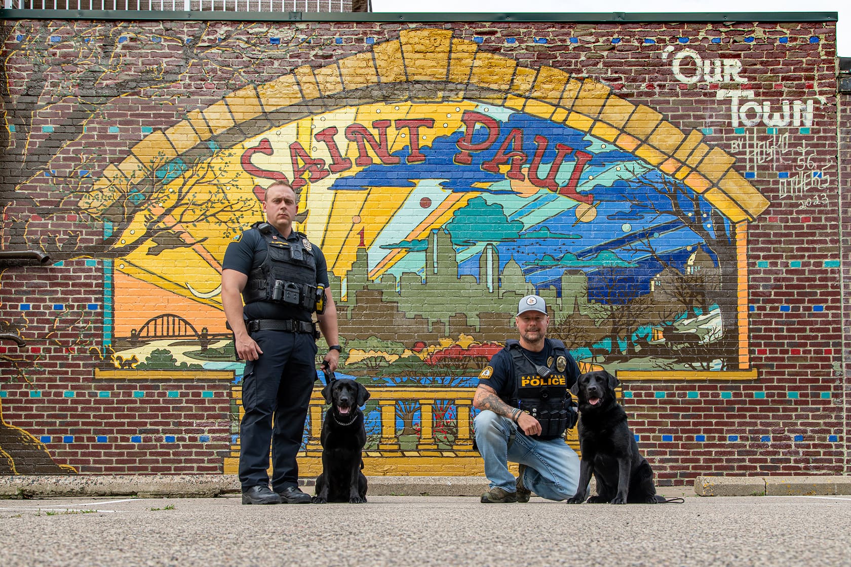Two police officers with black dogs posing in front of a colorful Saint Paul mural on a brick wall.