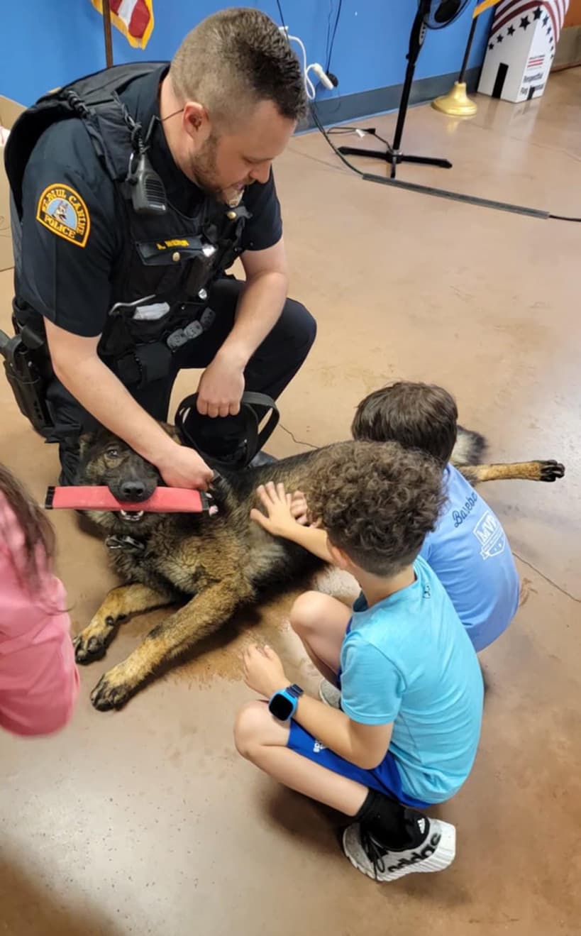 Police officer in blue uniform hugging a large black and tan German Shepherd dog indoors with colorful decorations on the wall behind.