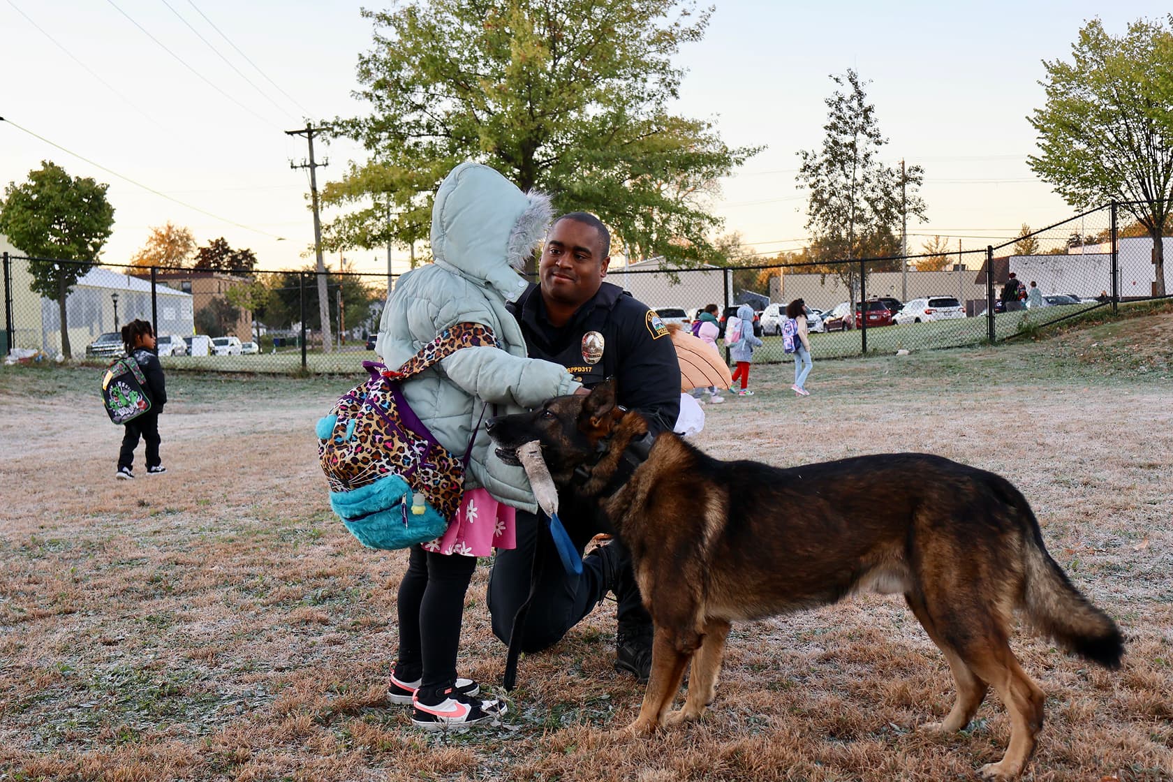 A police officer kneels to interact with a child wearing a teal jacket and carrying a leopard print backpack, while a large brown and black police dog stands nearby holding a toy.