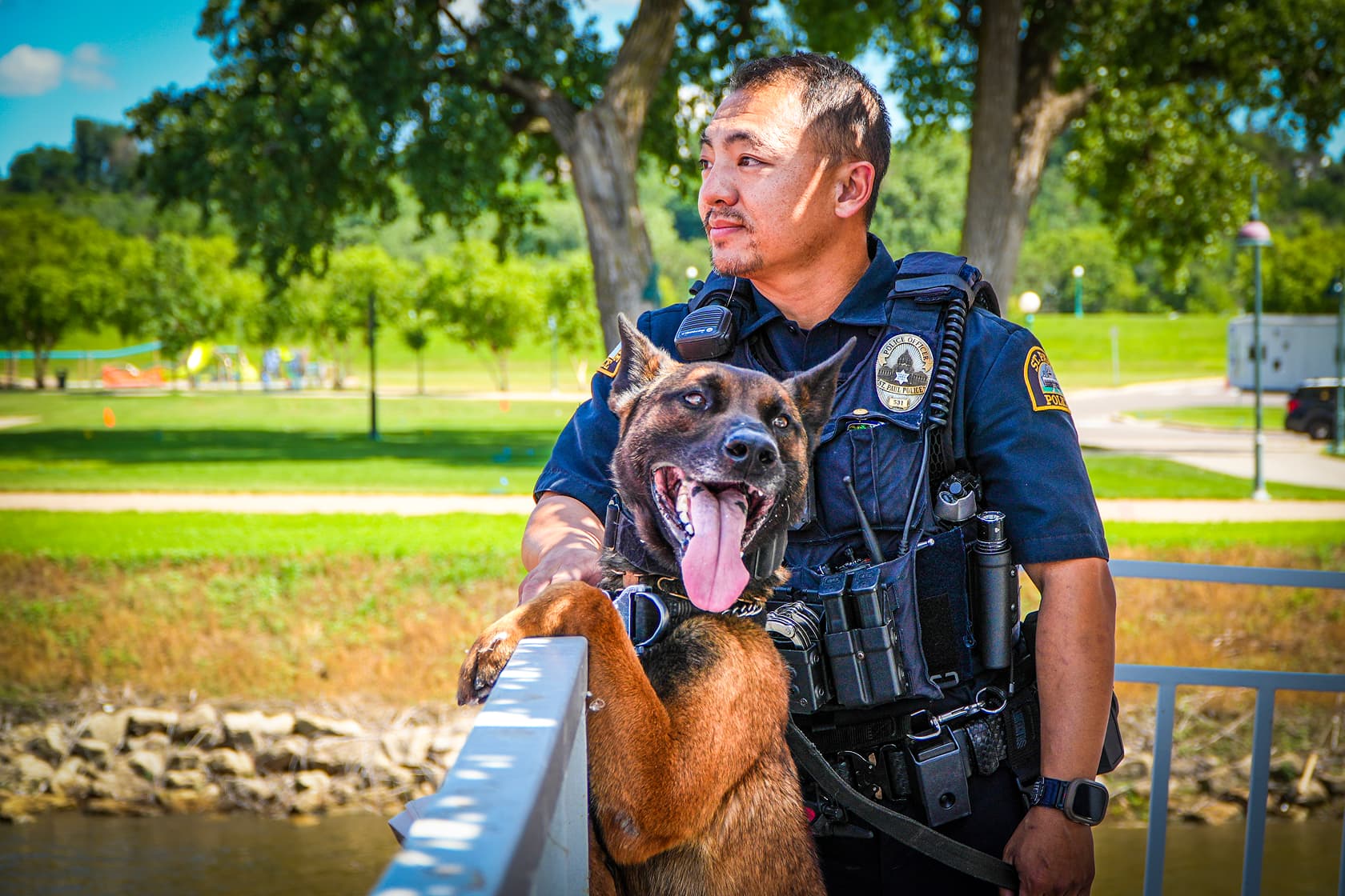 Police officer standing outdoors with a German shepherd police dog resting its front legs on a railing and panting.
