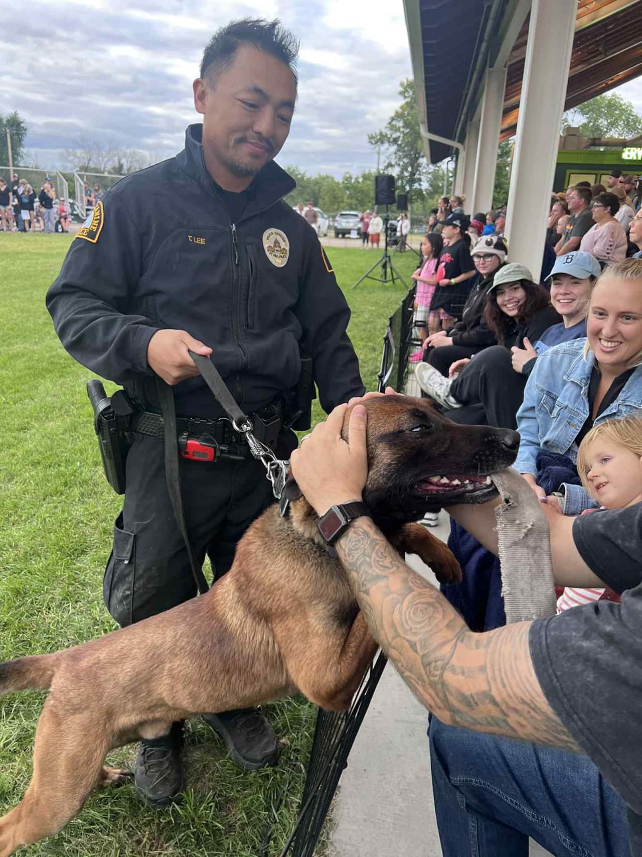 Police officer kneeling next to a hospital bed petting a German Shepherd, beside a smiling patient with medical electrodes on their forehead.