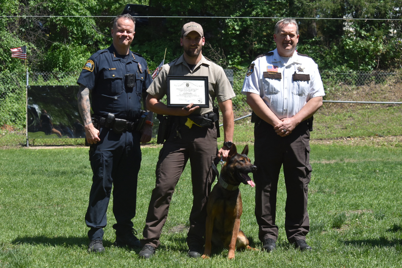 Three law enforcement officers stand on grass, center holds a certificate with a police dog sitting in front.