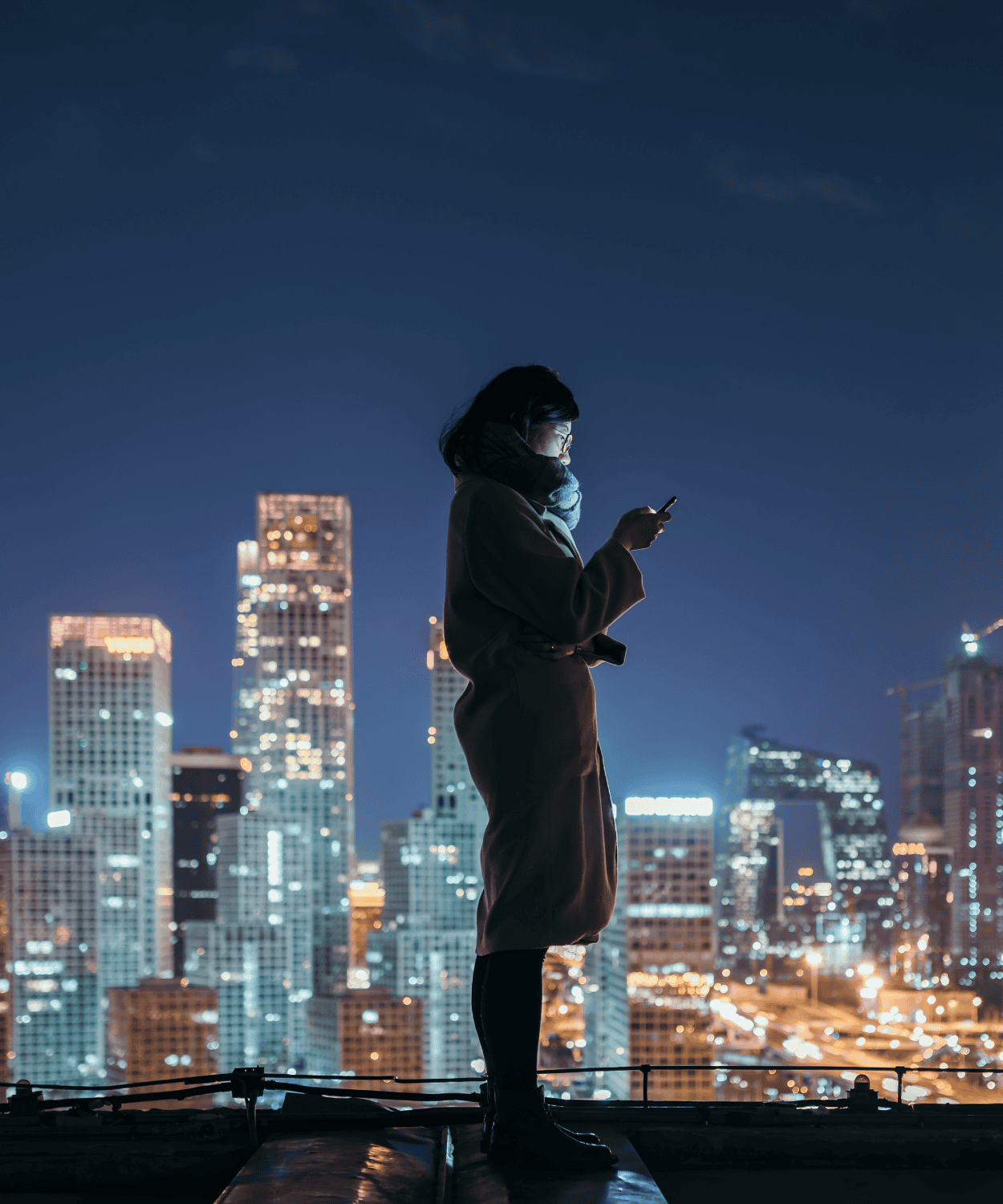 Silhouette of a person standing outdoors at night, looking at a phone, with illuminated skyscrapers and city lights in the background.