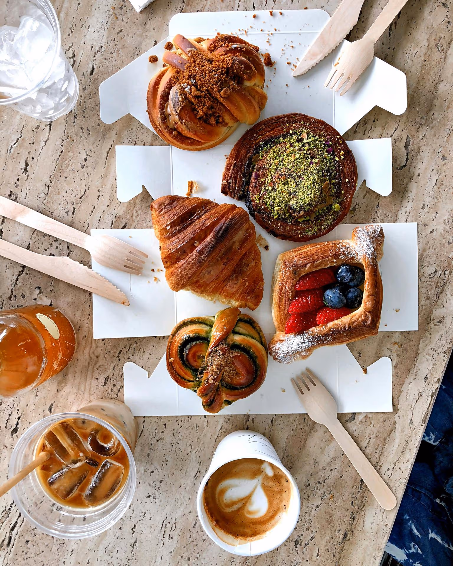 Aerial view of assorted pastries including a croissant and berry tart on a table with iced and hot coffee. Wooden utensils add a rustic touch.