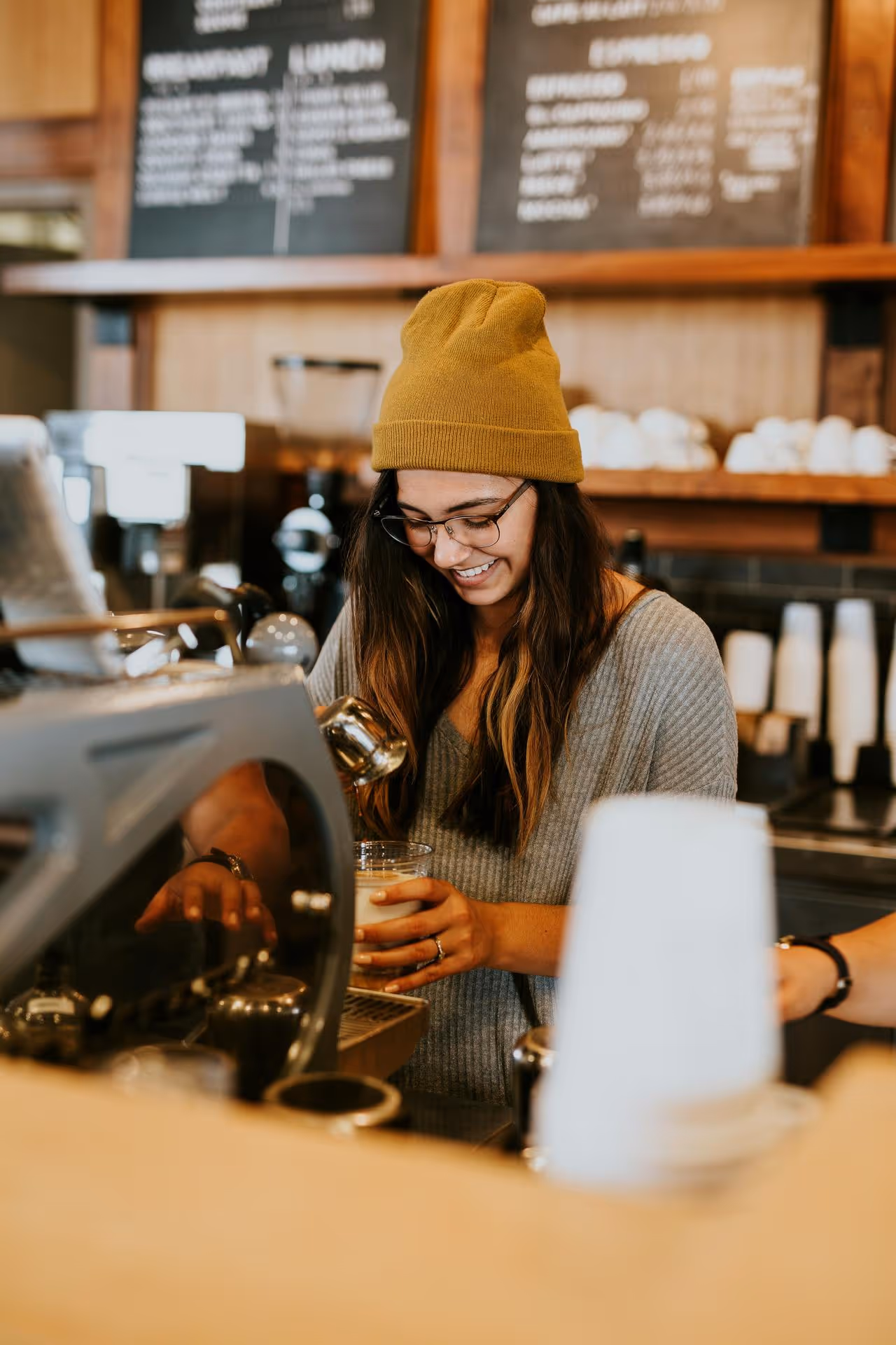 Barista wearing a mustard beanie and glasses smiles while making coffee at an espresso machine. Warm, cozy cafe atmosphere with blurred menu board.