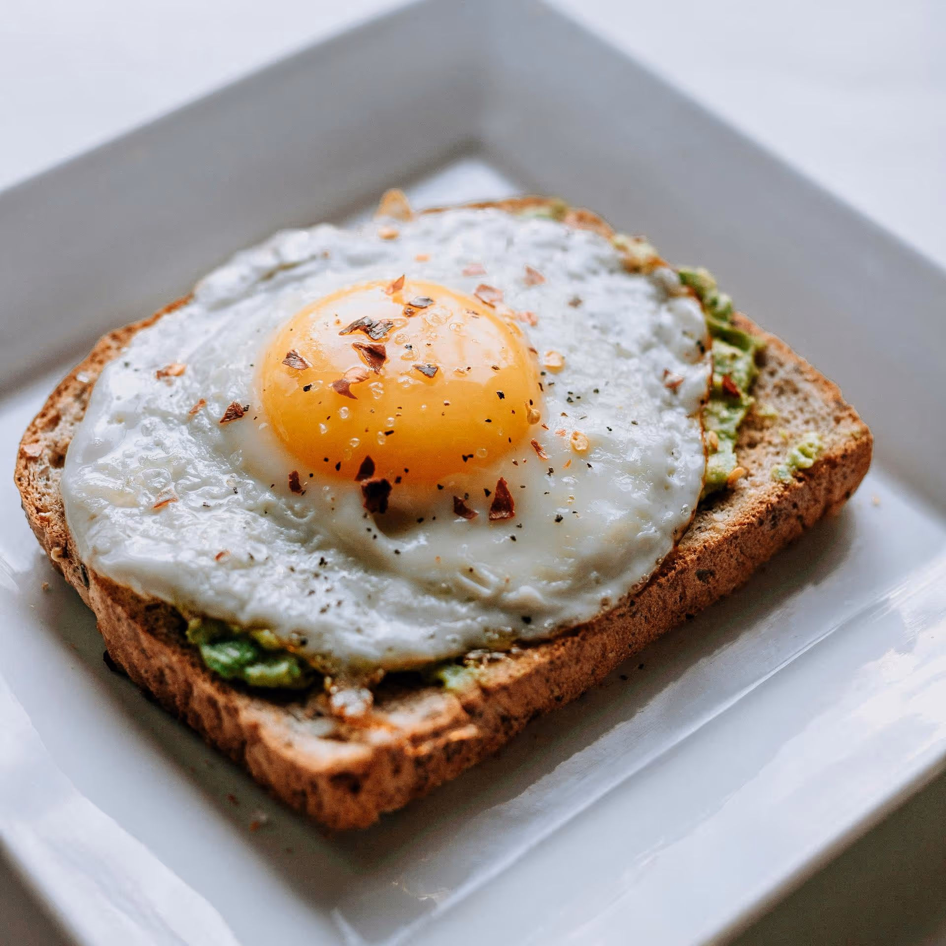 A slice of whole grain toast topped with a perfectly fried egg, featuring a bright yolk and sprinkled with chili flakes.