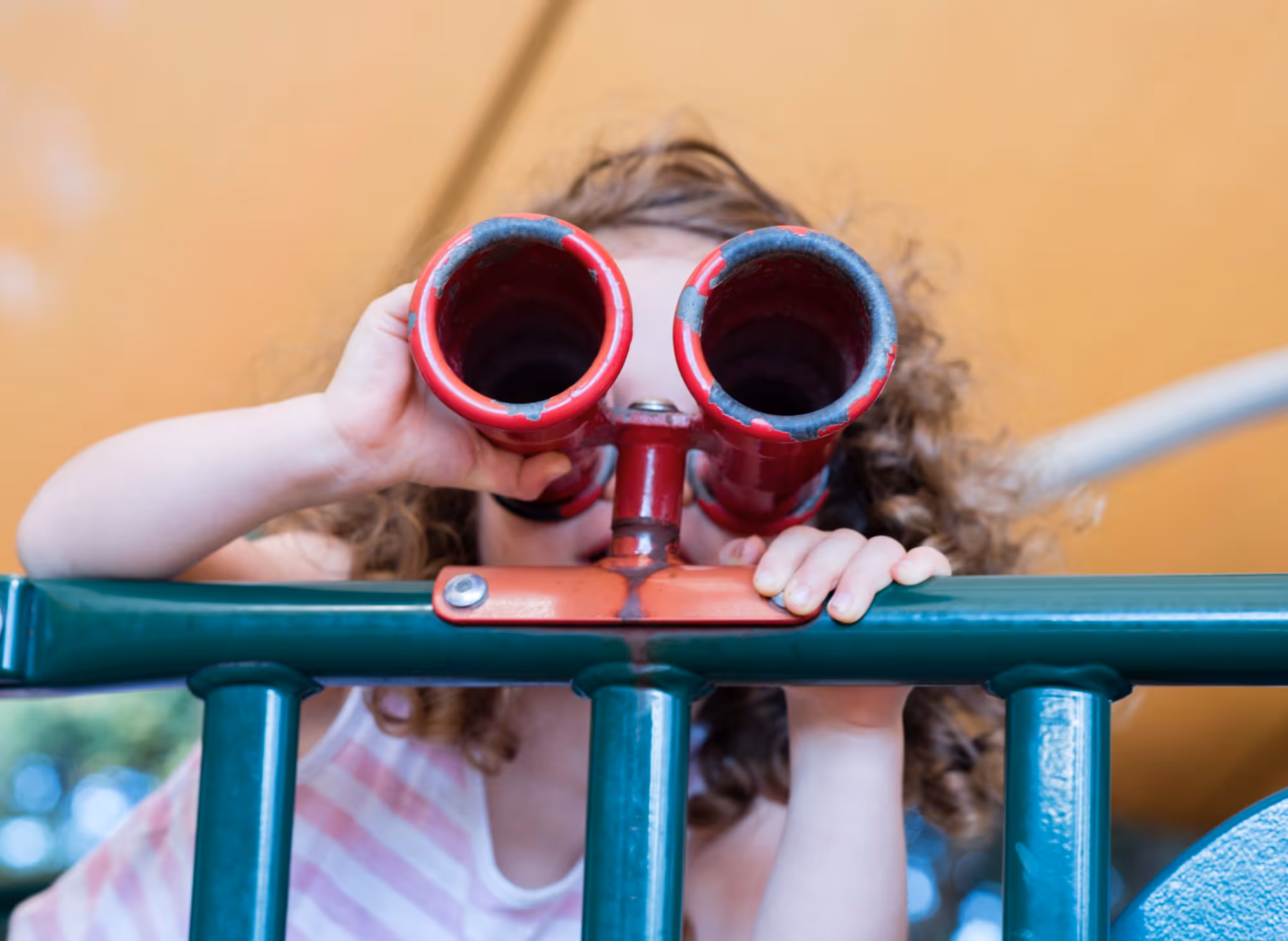 A young child looking through toy binoculars while leaning on playground equipment.