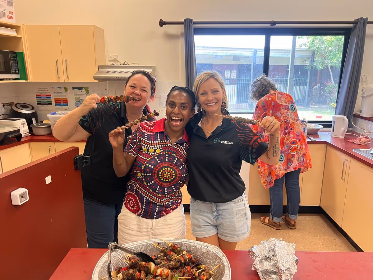 A group of women standing in a kitchen space, smiling and holding prepared food.