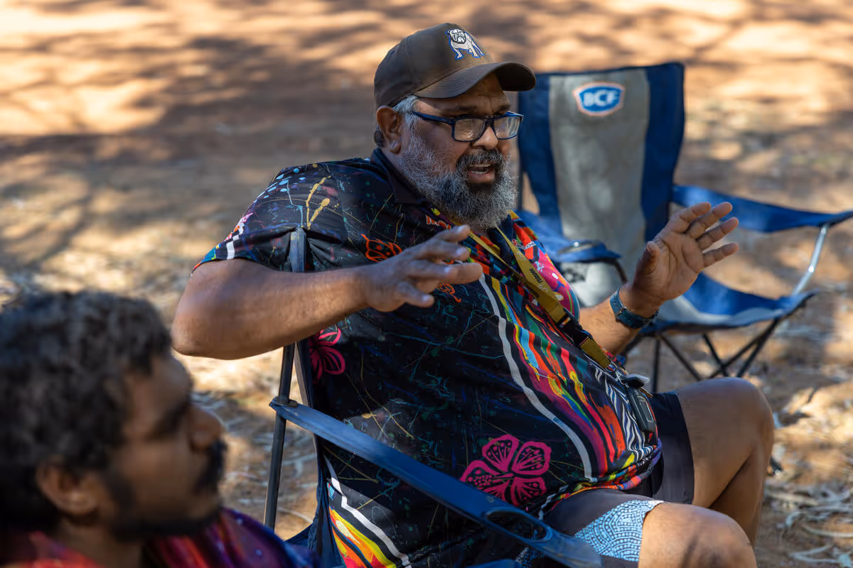 An adult speaking with children outdoors, seated in a casual community setting.