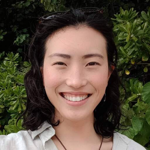 A photo of a Jenny smiling at the camera in front of some bushes, with sunnies on her head.
