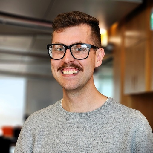 A photo of Jay smiling at the camera, wearing glasses, a grey sweater, and a moustache, in an office setting