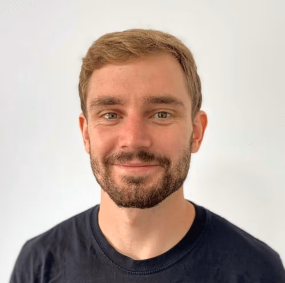 A photo of Josh smiling at the camera wearing a black tshirt and a beard against a light background