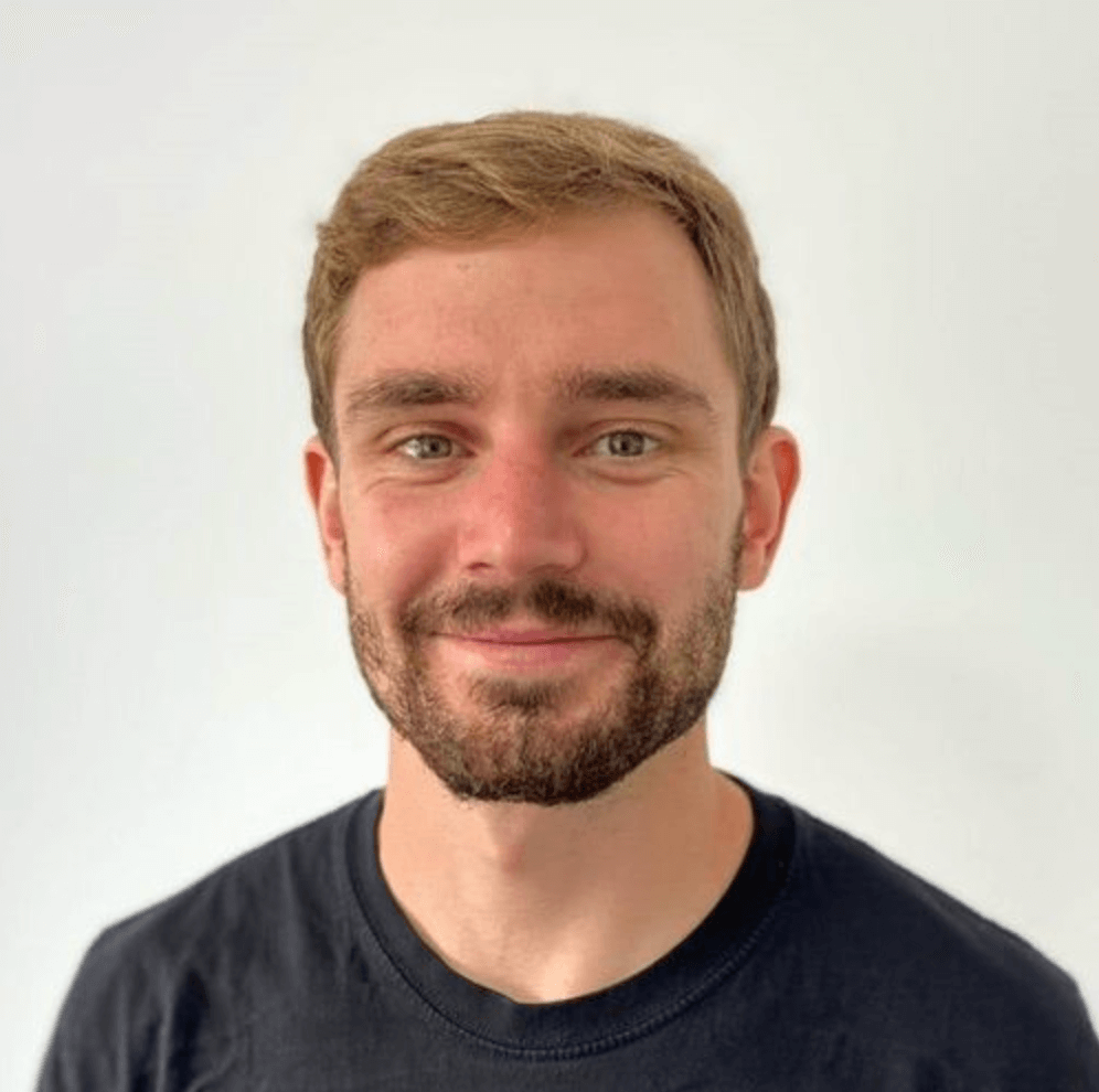 A photo of Josh smiling at the camera wearing a black tshirt and a beard against a light background