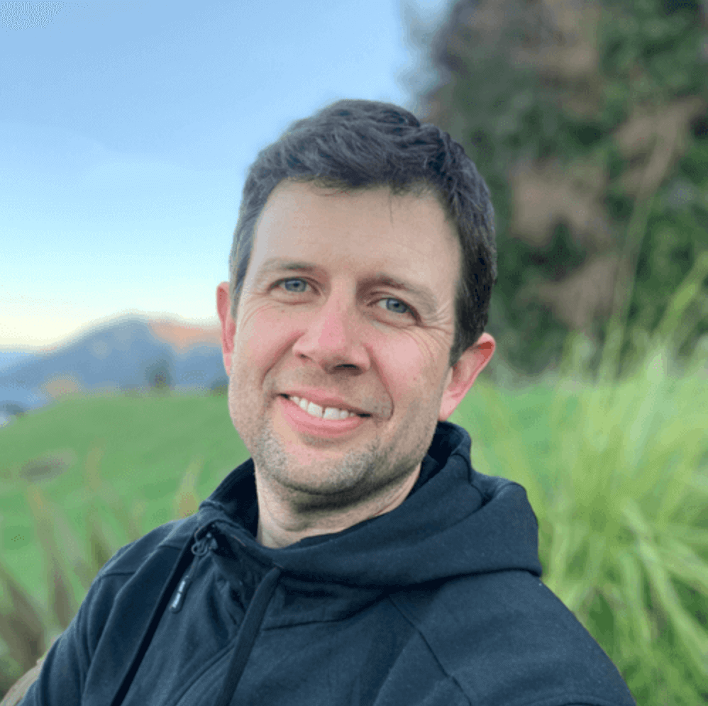 A photo of Steve smiling at the camera wearing a black hoodie against a grassy green background