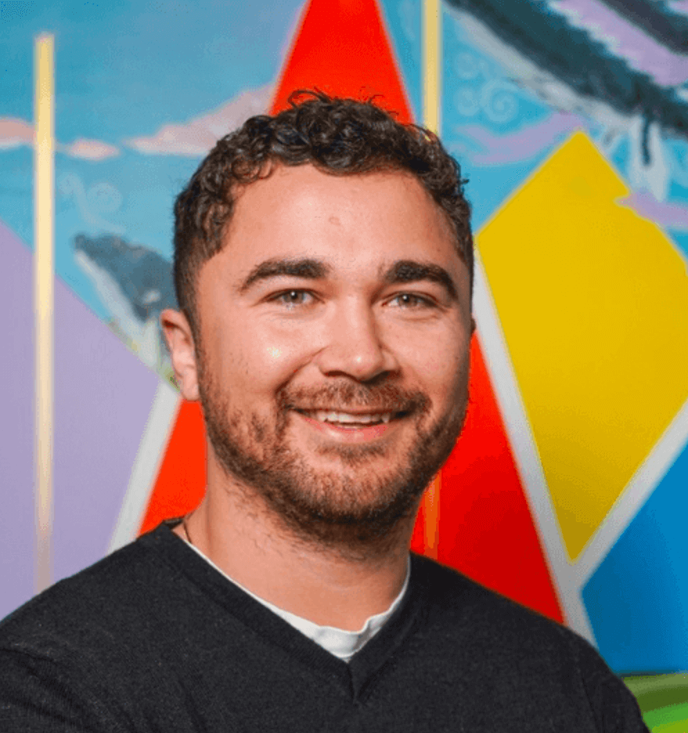 A photo of Mike smiling at the camera wearing a v-neck navy sweater against a bright colourful background