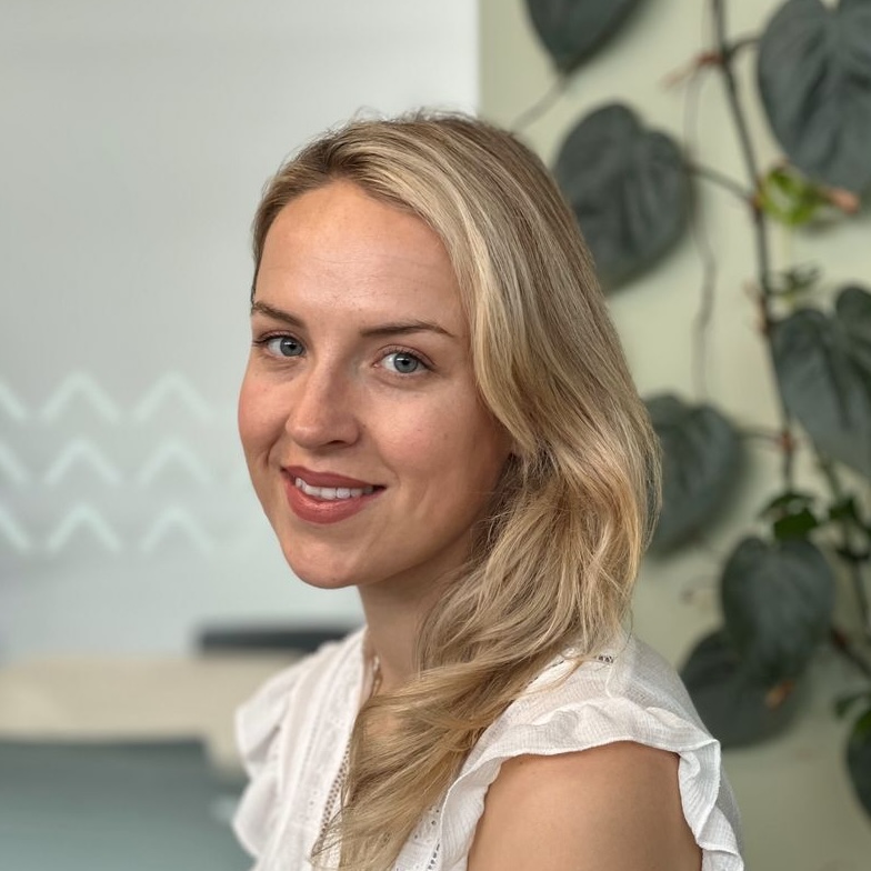A photo of Lowri smiling in a 3/4 angle towards the camera. She has blonde hair and a white shirt, with pothos leaves in the background