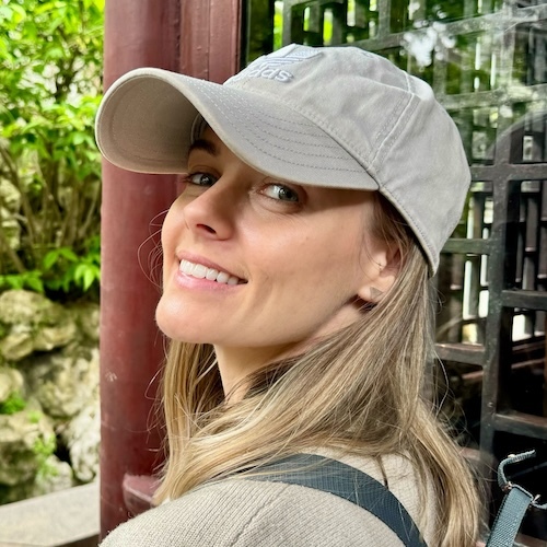 A photo of Jade, smiling over her shoulder with blonde hair wearing a grey baseball cap and a backpack, in front of a leafy background