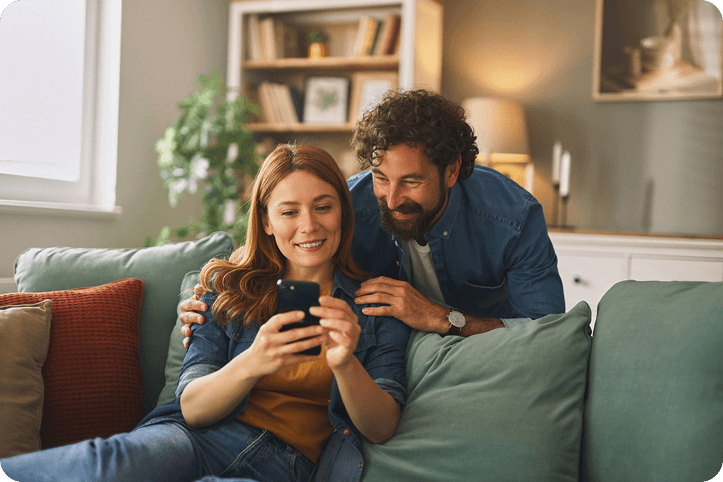 A smiling couple sitting on a couch looking at a smartphone, representing customers comparing Klarna vs Afterpay payment options.