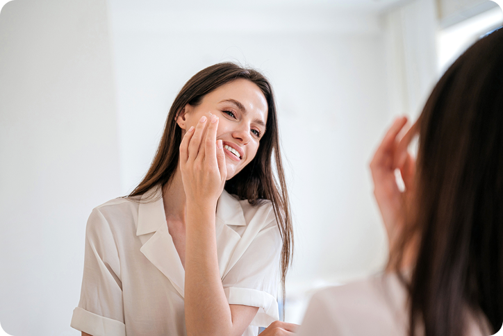 A smiling woman checks her skin in the mirror after being referred by a friend through a med spa referral program.