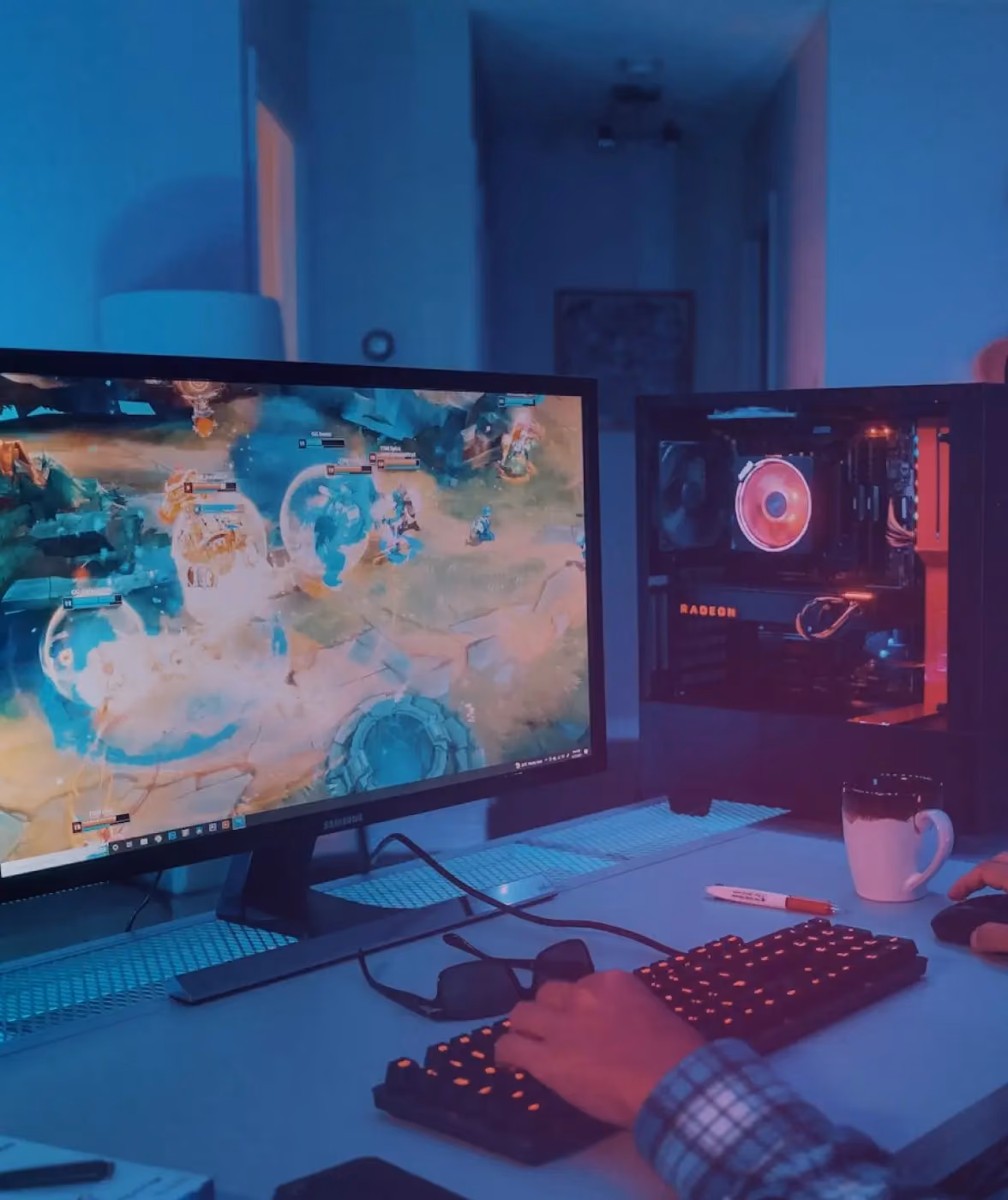 Person playing a multiplayer video game on a desktop computer with an illuminated mechanical keyboard and a gaming PC tower with red lighting on a desk in a dimly lit room.