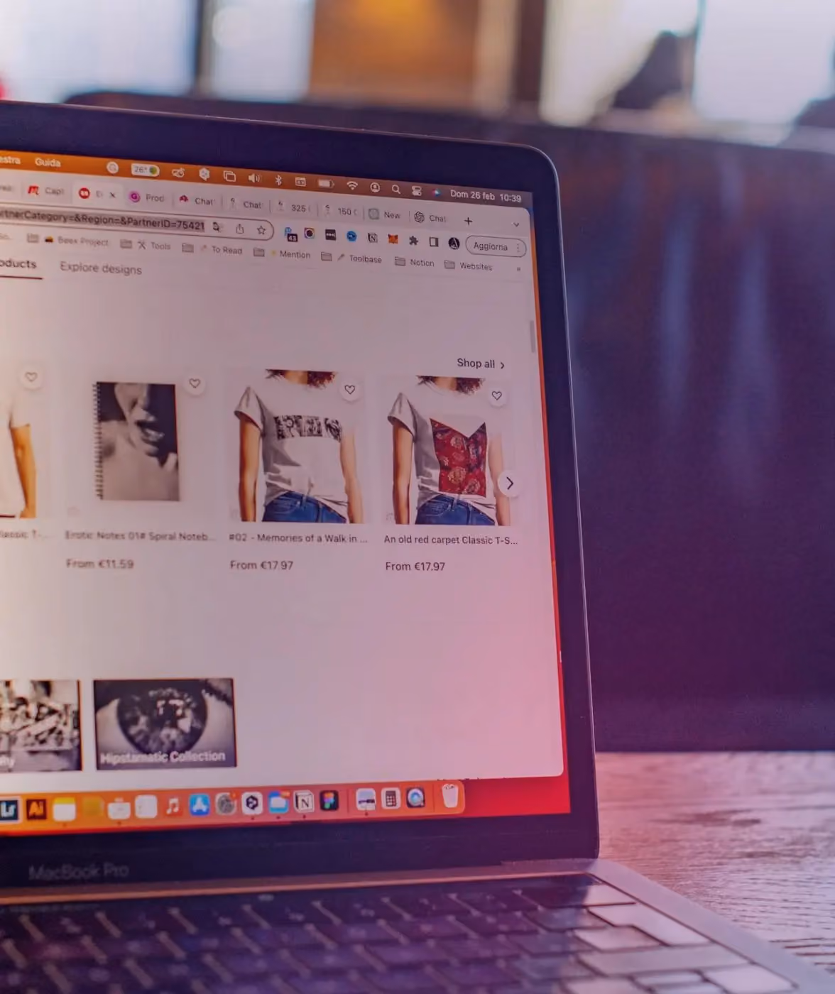 Close-up of a MacBook Pro laptop displaying an e-commerce website with product listings including t-shirts and a spiral notebook, each with prices in euros.