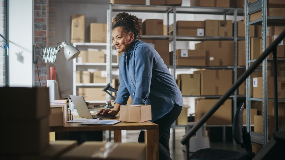 A woman standing in a warehouse-style office using a laptop, overlaid with buttons labeled “Purchase Orders,” “Approval Workflows,” “Invoice Processing,” and “Supplier Payments”