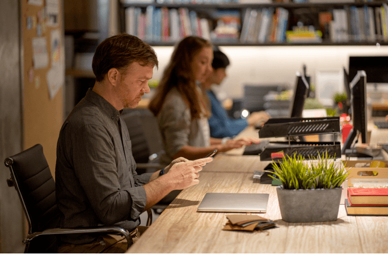 Office workers seated at a long shared desk using computers and mobile devices working on accounts payable automation software in a modern workspace with bookshelves and plants in the background.
