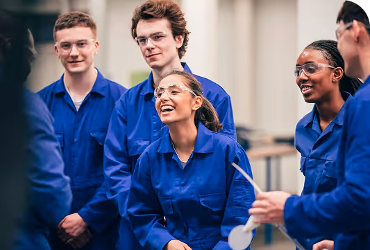 A group of people in what looks like a practical lab of some kind. They all wear blue vests and safety goggles and seem to be happy.
