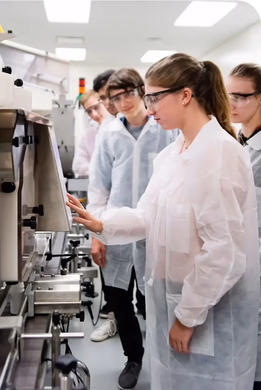 A group of people in what looks like a practical lab of some kind. They all wear clear white vests and safety goggles and seem to be focused.