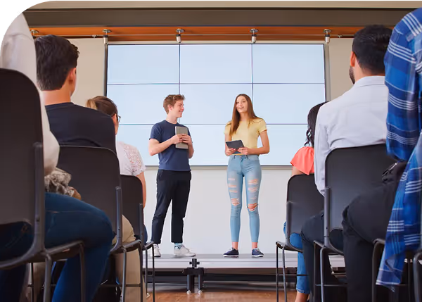 Two people present something on a small  stage resembling a teaching room.