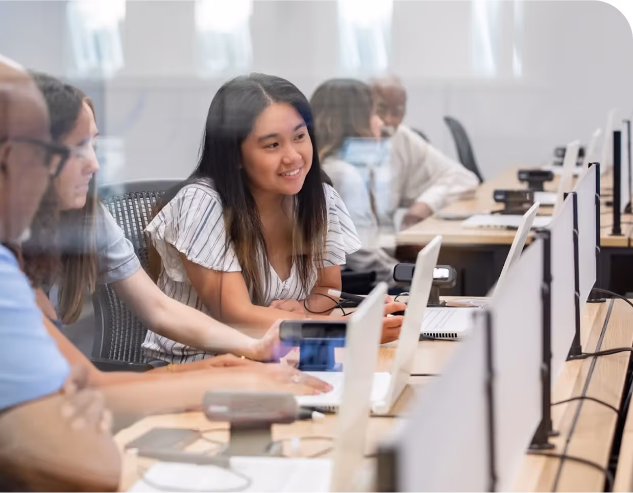 A group of people in desks, they each have a laptop and are in a row, apparently discussing some idea related to what's likely a class.