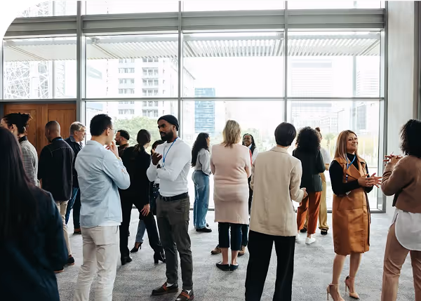 A group of people talk among each other in a big and tall room with large windows in the background.