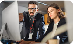 Two people work on a PC, one of them points at the monitor while the other one looks.