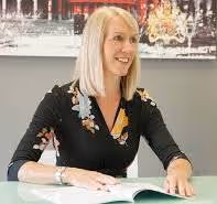 A woman with blonde hair smiles while opening a book at a desk.