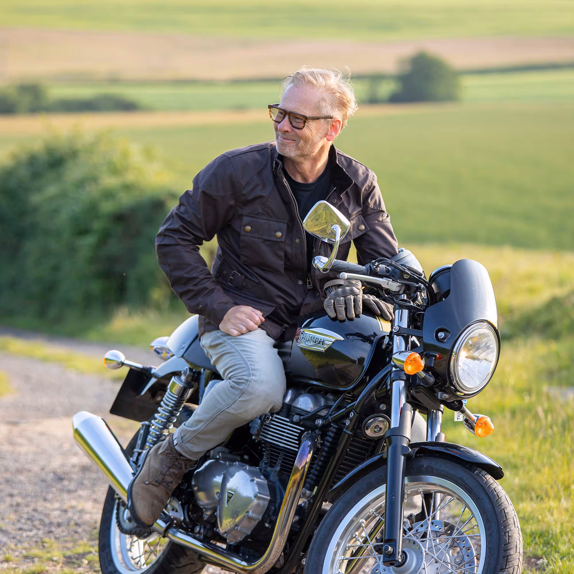 A person sits on a parked motorcycle, looking to the side, in a rural outdoor setting.
