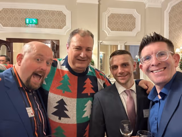 Four men in suits and festive sweater smiling at a gathering, holding drinks.