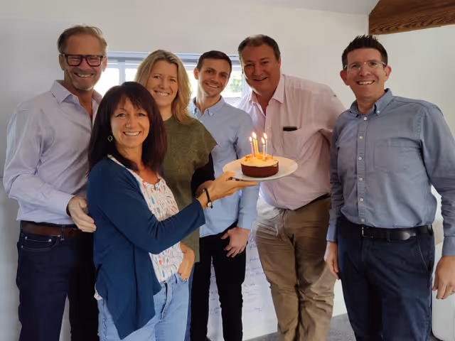 A woman holding a cake with candles stands with five smiling people indoors.