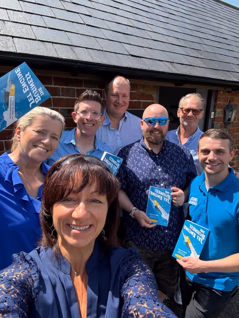A group of seven people smiling, holding books, posing for a selfie outside a brick building.