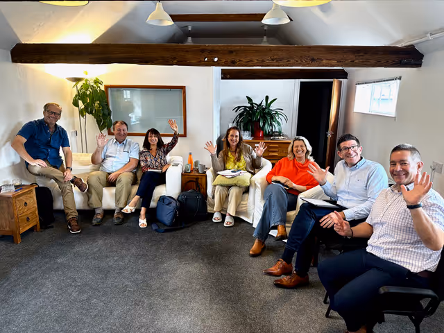 Seven people sitting in a room, smiling and waving towards the camera.