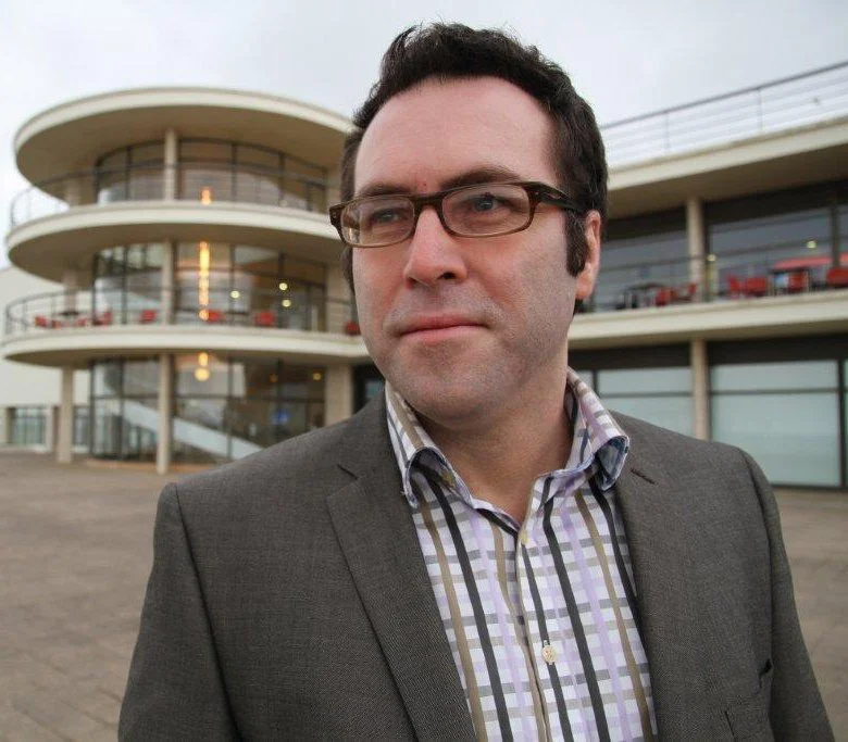 A man wearing glasses and a suit jacket stands in front of a modern circular building.