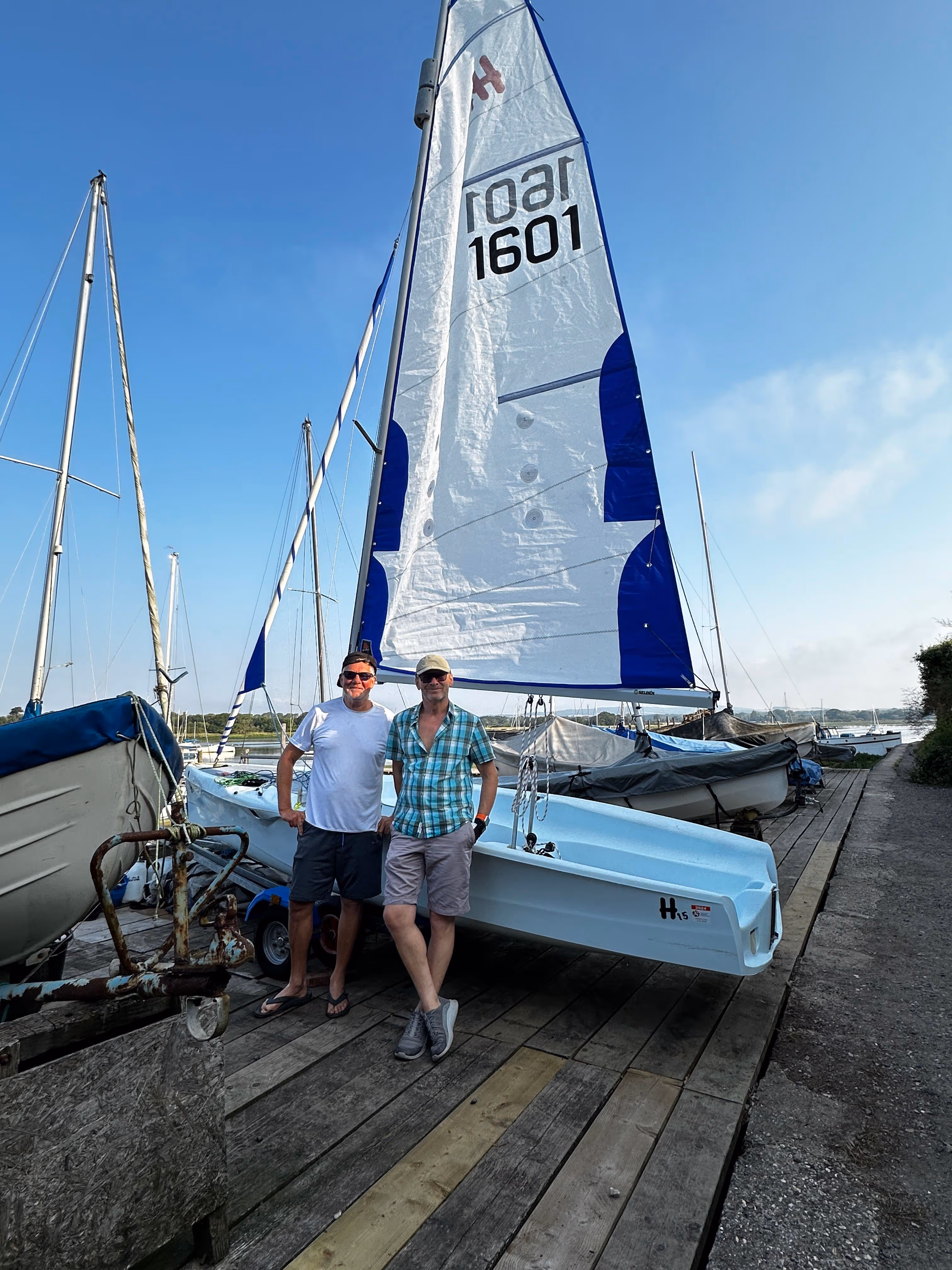 Two people standing in front of a parked sailboat on a wooden dock.