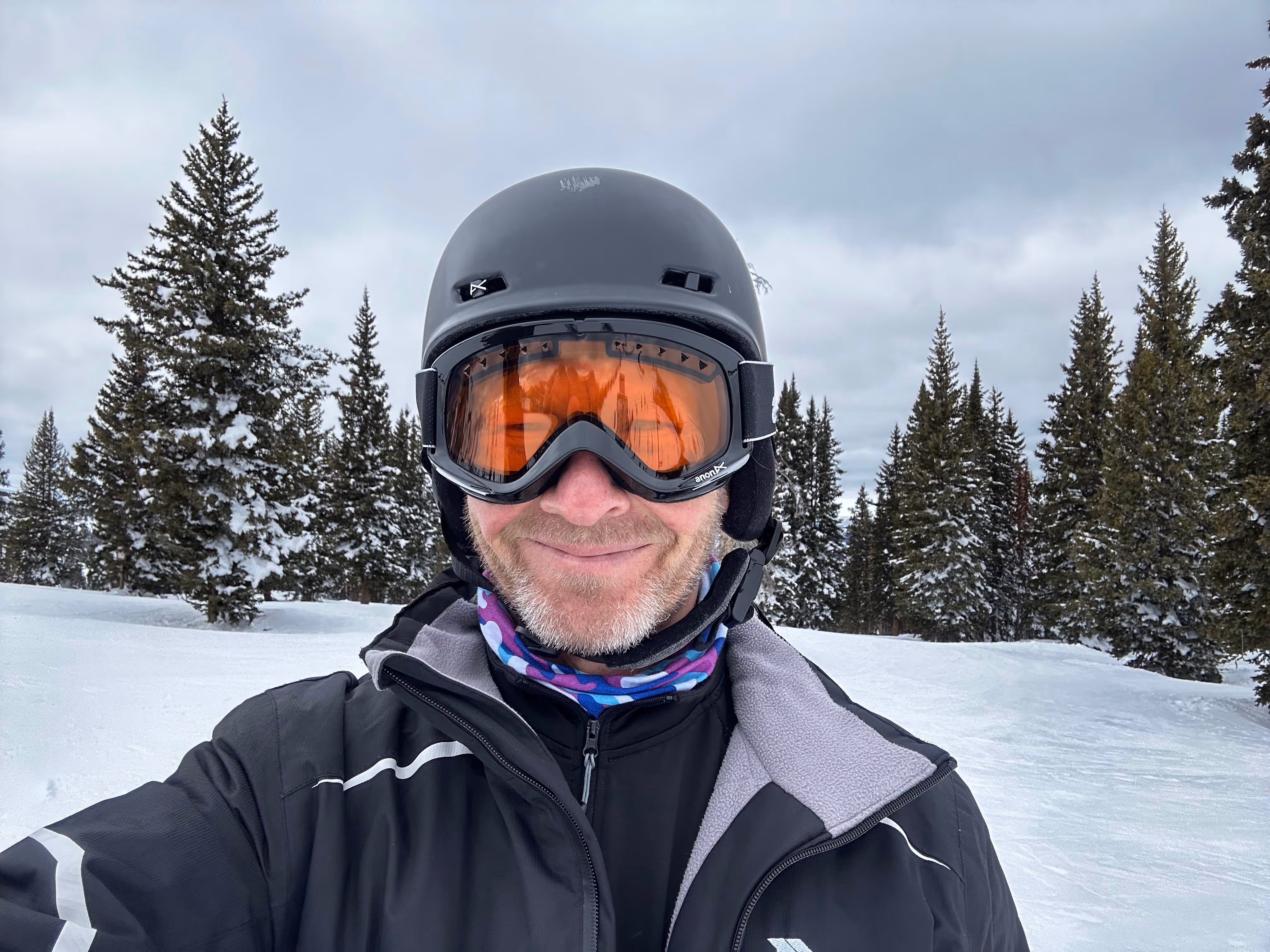 A person wearing ski gear and goggles smiles while standing in a snowy area with trees.
