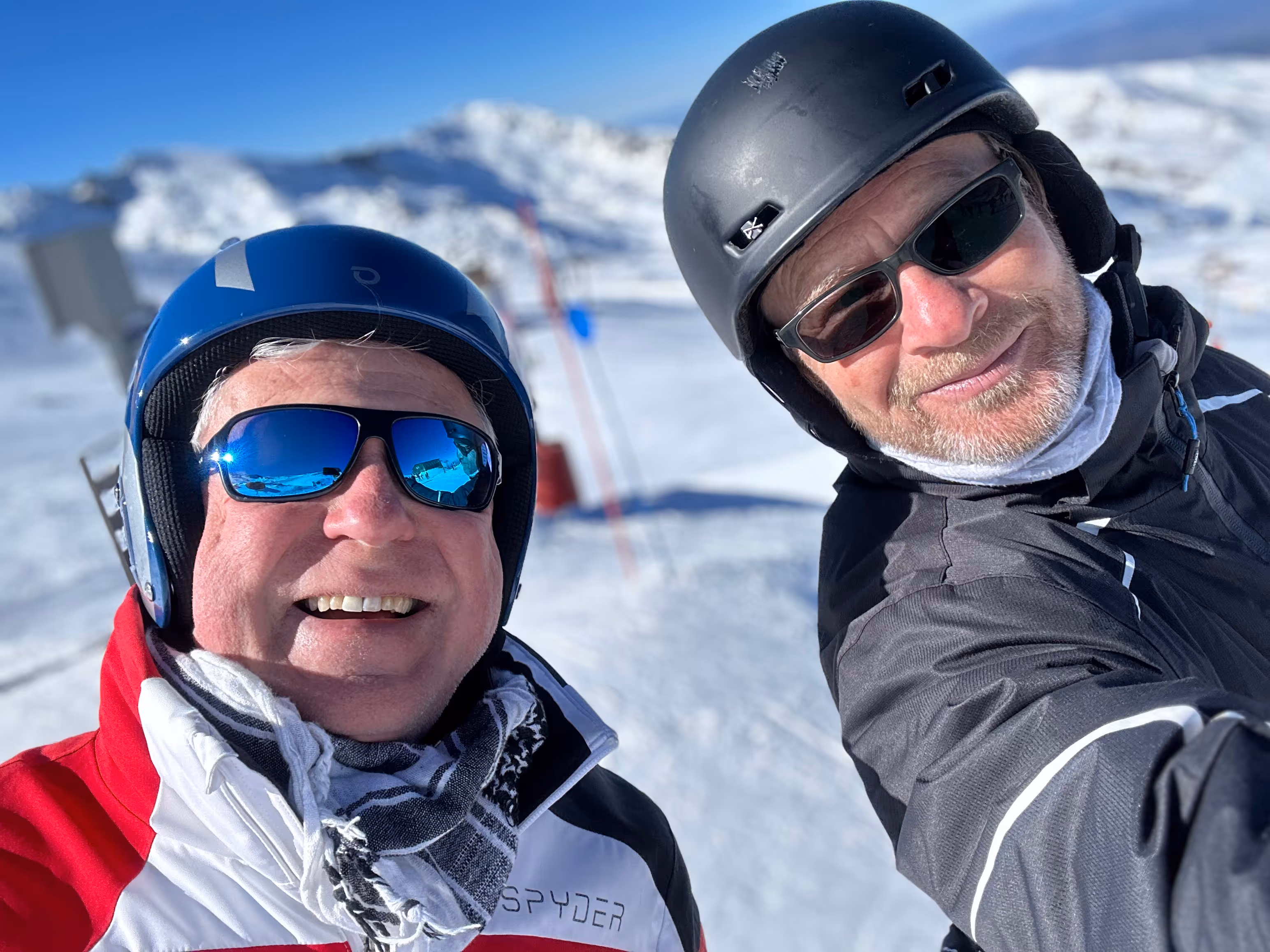 Two people wearing helmets and ski gear smile for a selfie in a snowy mountain setting.
